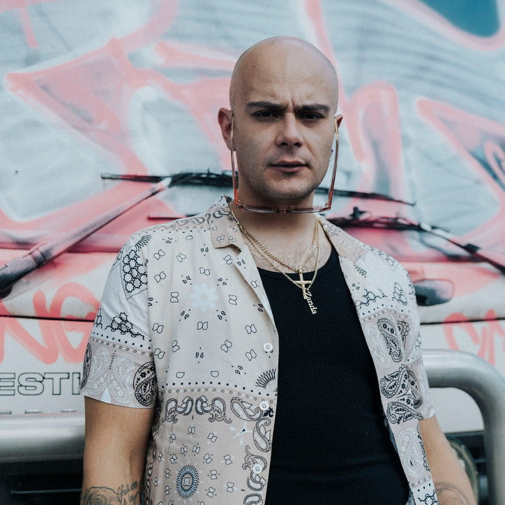 Man with tattoos wearing a patterned shirt and black tank top in front of a graffiti-covered vehicle.